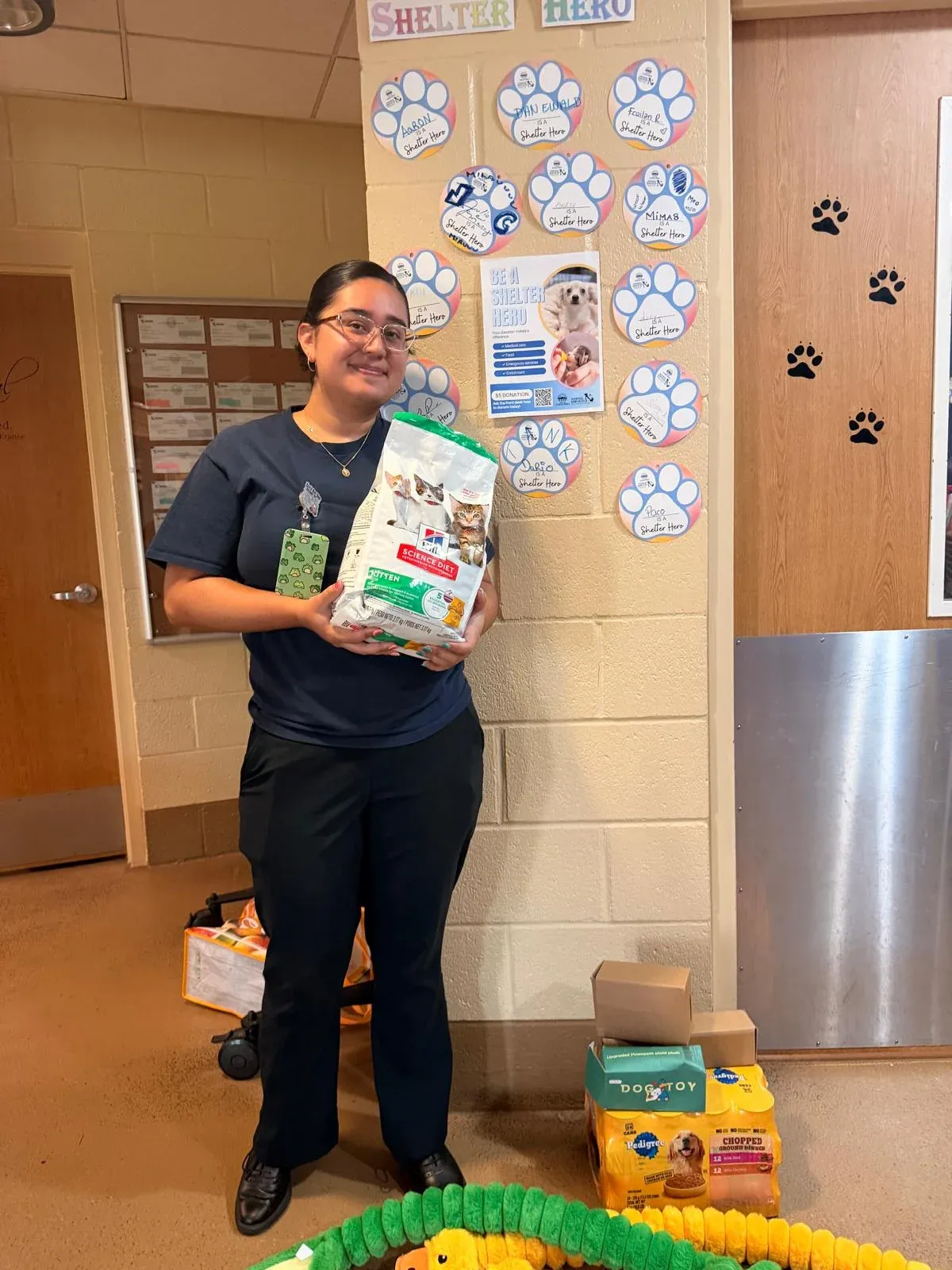 A Chula Vista Animal Services staff member holding a donated bag of pet food, with boxes of food, treats and toys on the floor beside her.